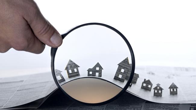 Hand holds the magnifying glass in front of an open newspaper with paper houses. That could mean rent, search, purchase real estate, property investment generic