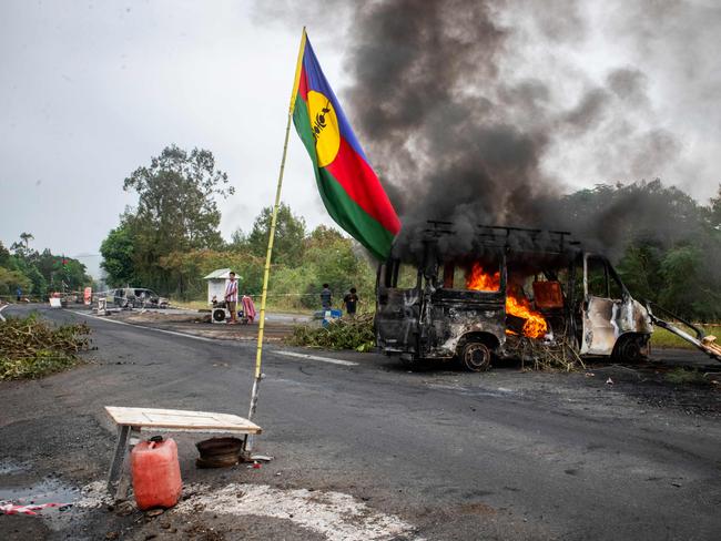 TOPSHOT - This photograph shows a Kanak flag waving next to a burning vehicle at an independantist roadblock at La Tamoa, in the commune of Paita, France's Pacific territory of New Caledonia on May 19, 2024. French forces smashed through about 60 road blocks to clear the way from conflict-stricken New Caledonia's capital to the airport but have still not reopened the route, a top government official said on May 19, 2024. (Photo by Delphine Mayeur / AFP)