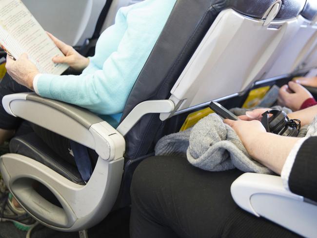 In this real situation a row of unidentifiable passengers are sitting in their seats on an airplane. The seating is tight. Their legs are touching the seats in front of them.