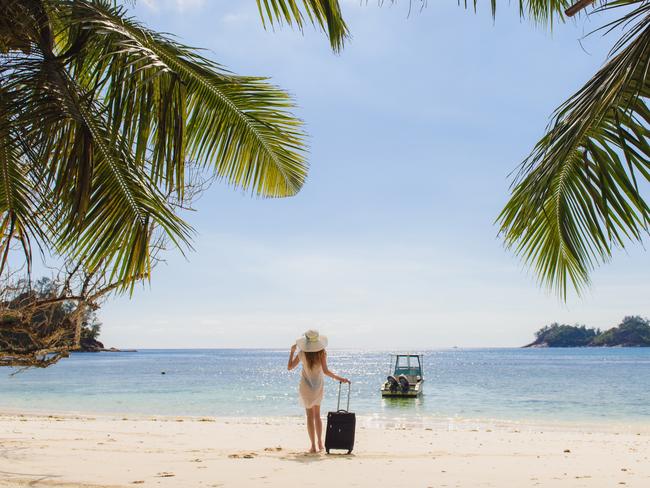 ESCAPE: Young woman with hat and suitcase on the beach. Transparent clothes, bathing suit. Palm fronds framed photo. Picture: Istock