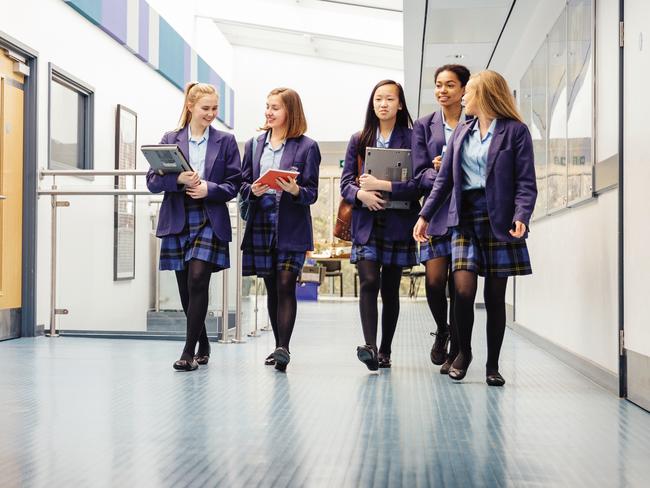 Group of teenage girls are walking down the school hall with books and laptops in their arms. They are talking and laughing as they walk and there is a female teacher using the printer.