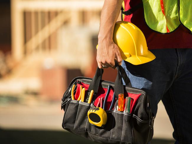 A construction worker busy working at a job site. He holds a tool box full of tools and a hard hat. Framed house, building in background. He is wearing a safety vest.