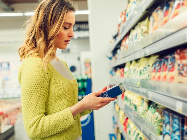 Young smiling woman groceries shopping in local supermarket. She is standing, holding box and reading nutrition facts.