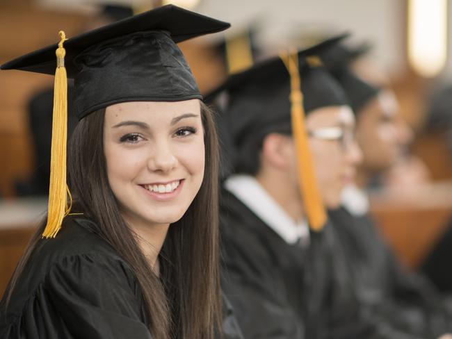 A multi-ethnic group of college graduates are sitting together in a lecture hall and are listening to their professor. They are wearing a cap and gown and one woman is smiling and looking at the camera. Photo: iStock