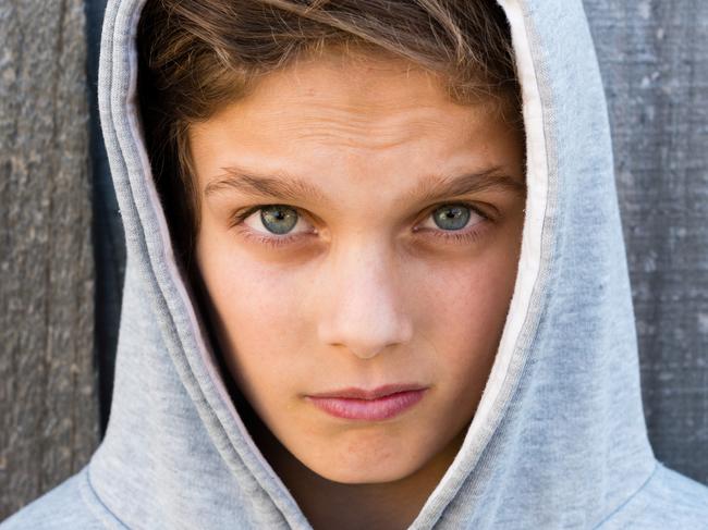 Portrait of beautiful teenage boy in hoodie with aggressive expression on his face against grey wooden fence