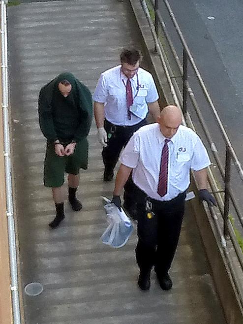 Chad Badcock in green escorted by security officers.                         <a class="capi-image" capiId="387bd8f062697464c5f7dd1aa9726918"></a>