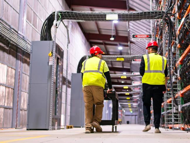 Two technicians wearing PPE walking past racks of equipment in a data center for cryptocurrency mining, cloud services and AI computing in a large, temperature controlled warehouse in a remote location in Stutsman County, North Dakota. One of them is pushing a cart.