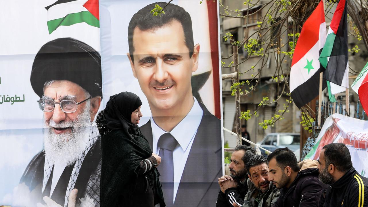 Portraits of Syria's President Bashar al-Assad (R) and Iran's supreme leader Ayatollah Ali Khamenei stand as Palestinians sit at the entrance of the Yarmuk camp for Palestinian refugees, south of Damascus, during a delivery of humanitarian aid provided by Iran as part of the Muslim holy fasting month of Ramadan on March 26, 2024. (Photo by LOUAI BESHARA / AFP)