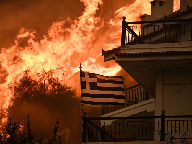 (FILES) A Greek flag flutters in the wind during a wildfire in Chasia in the outskirts of Athens on August 22, 2023. The summer of 2023 (June-July-August) saw the highest average global temperatures ever measured, announced on September 6, 2023 the European observatory Copernicus, according to whom 2023 will probably be the hottest year in history. (Photo by Angelos Tzortzinis / AFP)