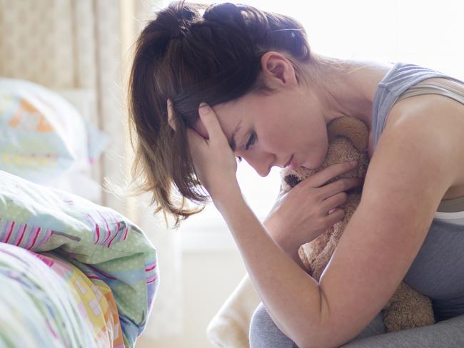 A grieving mother sits in her child's bedroom holding their toy in her arms while resting her head in her hands. The child's bed can be seen in the background untouched from when they were last in it.