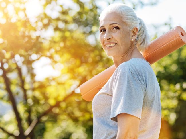 Stay active. Cheerful smiling aged woman holding yoga carpet on the shoulder and looking in the camera while standing in the park. Healthy baby boomers and seniors, generic retirement