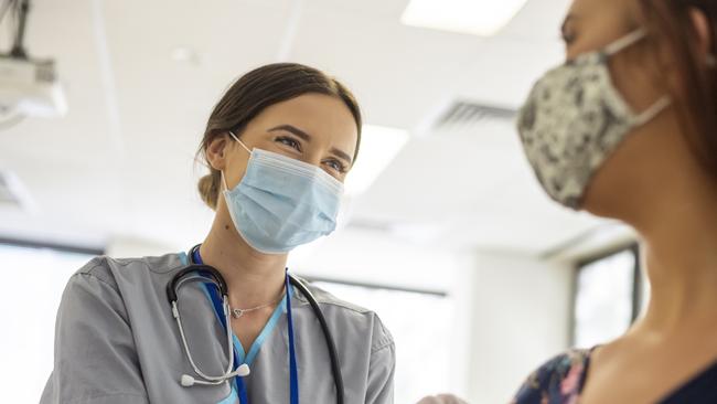 Shot focused on a kind looking nurse wearing scrubs, a stethoscope, white rubber gloves and a protective face mask. She is smiling at the nervous patient with her eyes. The nurse is prepping the patients arm before she injects her with the COVID-19 vaccine.