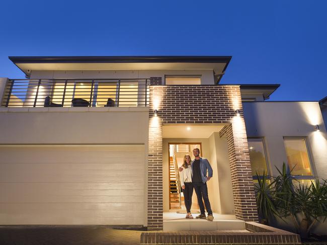 Couple standing in front of their new home. They are both wearing casual clothes and embracing. They are looking at the camera and smiling. The house is contemporary with a brick facade, driveway, balcony and a green lawn. The open front door is also visible. It is evening, twilight or night. Copy space