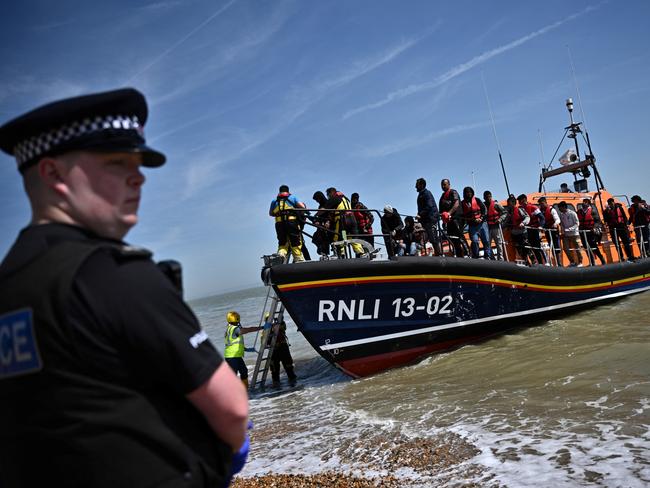 A British police officer stands guard on the beach of Dungeness, on the southeast coast of England, on June 15, 2022, as Royal National Lifeboat Institution's (RNLI) members of staff help migrants to disembark from one of their lifeboat after they were picked up at sea while attempting to cross the English Channel. Furious Conservatives called on Britain's government on June 15, 2022 to abandon a European human rights pact after a judge dramatically blocked its plan to fly asylum-seekers to Rwanda. Under the UK's agreement with Rwanda, all migrants arriving illegally in Britain are liable to be sent to the East African nation thousands of miles away for processing and settlement. The government, after arguing that Brexit would lead to tighter borders, says the plan is needed to deter record numbers of migrants from making the perilous Channel crossing from northern France. (Photo by Ben Stansall / AFP)