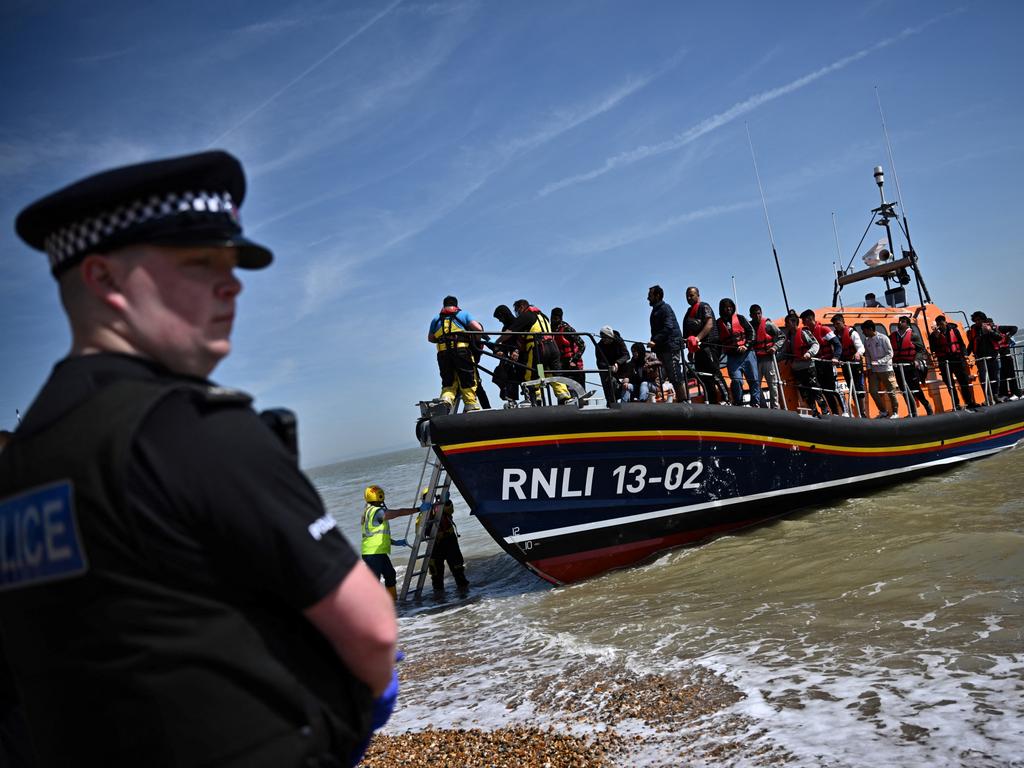 A British police officer stands guard on the beach of Dungeness, on the southeast coast of England, on June 15, 2022, as Royal National Lifeboat Institution's (RNLI) members of staff help migrants to disembark from one of their lifeboat after they were picked up at sea while attempting to cross the English Channel. Furious Conservatives called on Britain's government on June 15, 2022 to abandon a European human rights pact after a judge dramatically blocked its plan to fly asylum-seekers to Rwanda. Under the UK's agreement with Rwanda, all migrants arriving illegally in Britain are liable to be sent to the East African nation thousands of miles away for processing and settlement. The government, after arguing that Brexit would lead to tighter borders, says the plan is needed to deter record numbers of migrants from making the perilous Channel crossing from northern France. (Photo by Ben Stansall / AFP)