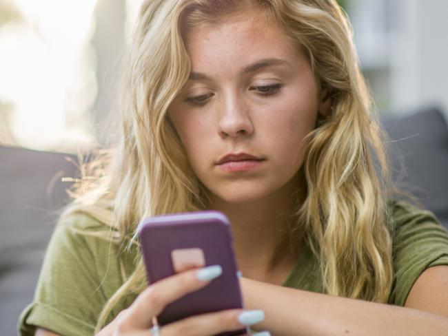 A young Caucasian woman is alone in her living room. She is sitting in front of the sofa and looking solemnly at her smartphone.