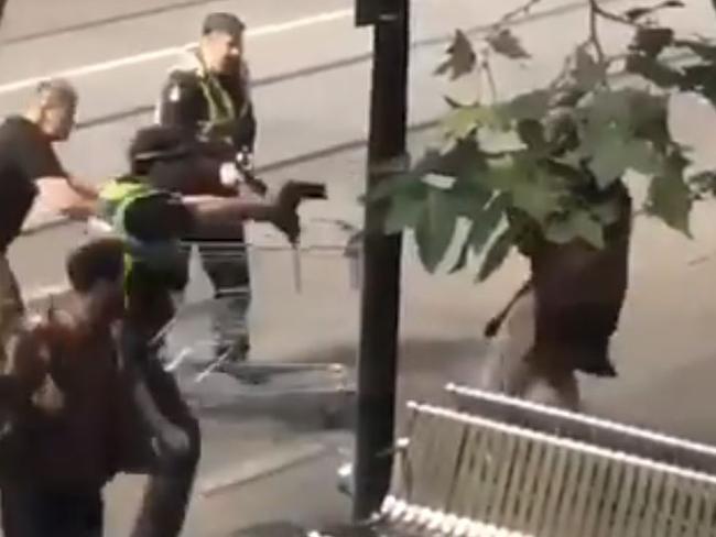 A policeman aims his gun at the man as a passer-by with a shopping trolley attempts to help.