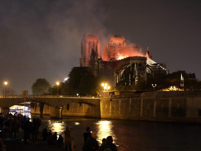 People stand on the banks of the River Seine as flames engulf the Notre-Dame Cathedral in Paris on April 15, 2019. - A colossal fire swept through the famed Notre-Dame Cathedral in central Paris on April 15, 2019, causing a spire to collapse and raising fears over the future of the nearly millenium old building and its precious artworks. The fire, which began in the early evening, sent flames and huge clouds of grey smoke billowing into the Paris sky as stunned Parisians and tourists watched on in sheer horror. (Photo by ludovic MARIN / AFP)