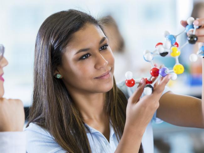 Teenage Hispanic female high school student is using plastic educational model toy molecules while studying in private school science class. Girl is holding study material while talking to teacher. Student is wearing a private school uniform.