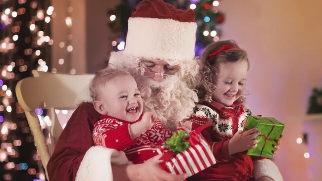 Santa Claus and children opening presents at fireplace. Kids and father in Santa costume and beard open Christmas gifts. Little girl helping with present sack. Family under Xmas tree at fire place.