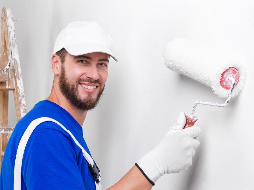 Portrait of handsome young smiling painter in white dungarees, blue t-shirt, cap and gloves painting a wall with paint roller, looking at camera