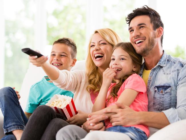 A happy family watches TV at home.  A dad in a denim shirt holds a little girl in a pink t-shirt.  A smiling mom holds a container of popcorn and directs a remote control toward a television.  A young boy wears a turquoise t-shirt and a pair of blue jeans.