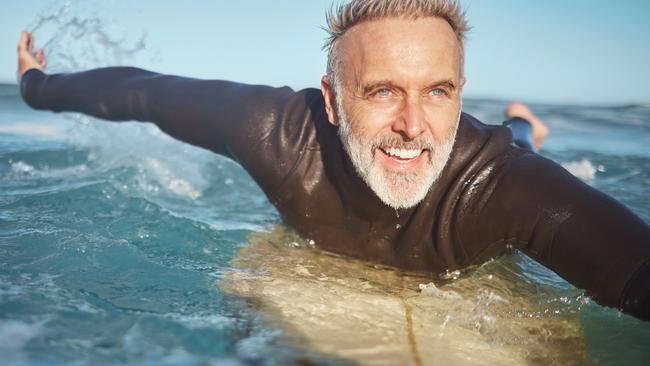 Beach, water and old man surfer swimming on a summer holiday vacation in retirement . Smile, ocean and senior surfing or body boarding enjoying a healthy exercise. Do the things you love to help your mental health.