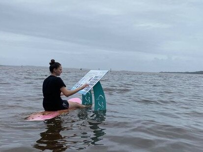 Camryn Chisholm inspecting an information sign on a surfboard. The sign is on a path around Tuggerah Lake which has been flooded by torrential rain. Picture: Morgan Chisholm