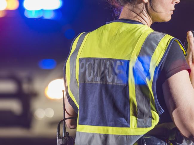 Rear view of a female police officer standing in the street at night, talking into her radio. Her patrol car is in the background with the emergency lights illuminated. She is wearing a yellow safety vest. Tablet generic