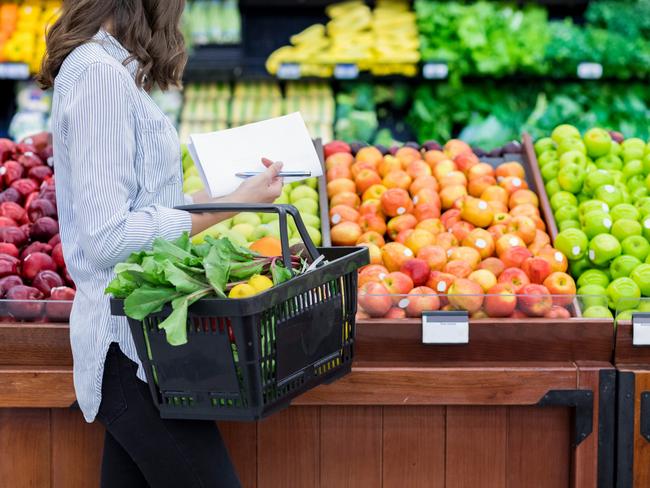 Young woman carries a shopping basket filled with fresh produce. She is shopping for fresh fruit and vegetables in a grocery store.