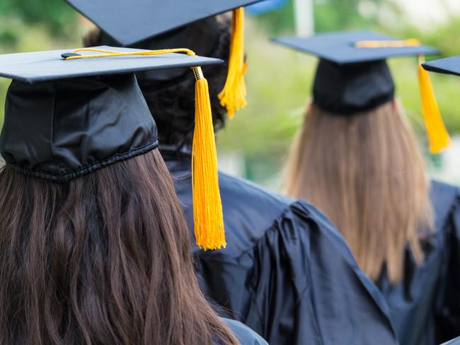 Group of students wearing graduation caps and gowns are walking on campus lawn. Young men and women are graduating from high school or college. Photo is of backs of students heads as they walk to receive their diplomas. Black caps have gold tassles.