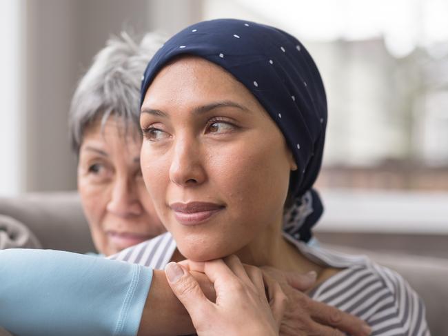 An ethnic woman wearing a headscarf and fighting cancer sits on the couch with her mother. She is in the foreground and her mom is behind her, with her arm wrapped around in an embrace, and they're both looking out the window in a quiet moment of contemplation.