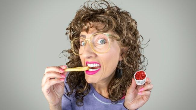 A fisheye image of a funny woman with big permed hair and glasses munching on a french fry.