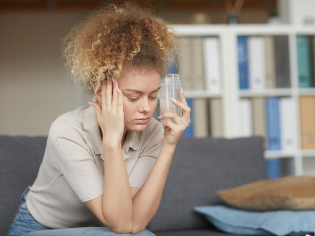 MENTAL HEALTH -  Young woman with curly hair sitting on sofa she has a headache and drinking medicine from it Picture: Istock