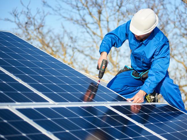 Male engineer in blue suit and protective helmet installing photovoltaic panel system using screwdriver. Professional electrician mounting solar module on roof. Alternative energy ecological concept.