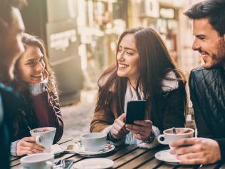 Laughing young people looking at phone and laughing while sitting in cafe outdoors, holding cups.