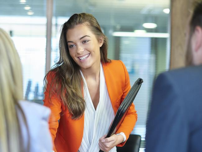 a young graduate walks into an interview room full of confidence and positivity energy . She is holding her cv and smiling at the interview panel before her. She is wearing blue trousers with an orange suit jacket , as she opens the door and strides in and shakes hands with a woman on the panel . in the foreground we can see the back of two of the panel , a man and a woman.