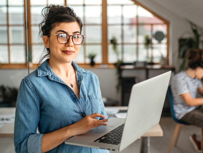 Portrait of young smiling woman in creative office, standing, holding laptop, looking at camera.