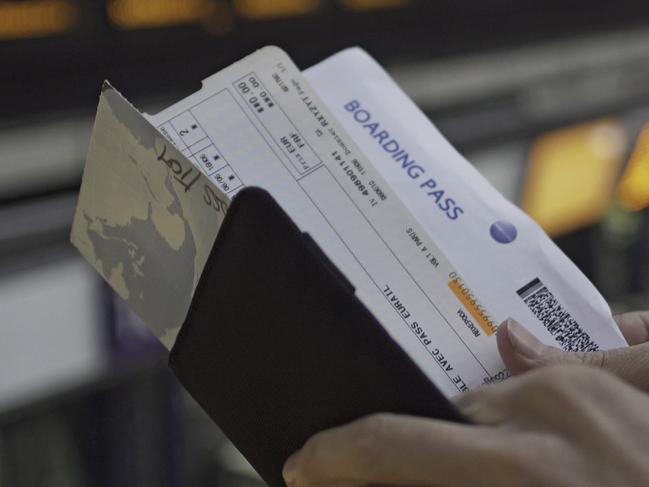 A pair of man's hands checks his travel documents and the time on his wristwatch. In the background is a large, out of focus, electronic departures board.