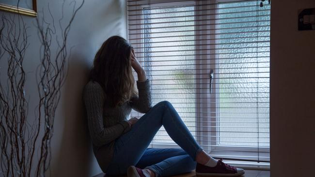 DTR914 Young female with long hair wearing jeans, hand on head in despair, sitting in darkness by a window at home. Side viewpoint.