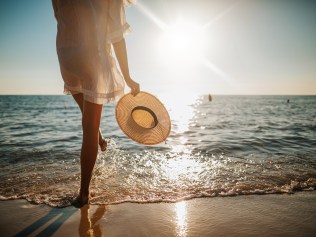 Close-up of young woman in white sun dress and with hat in hand walking alone on sandy beach at summer sunset, splashing water in sea shallow. Picture: iStock