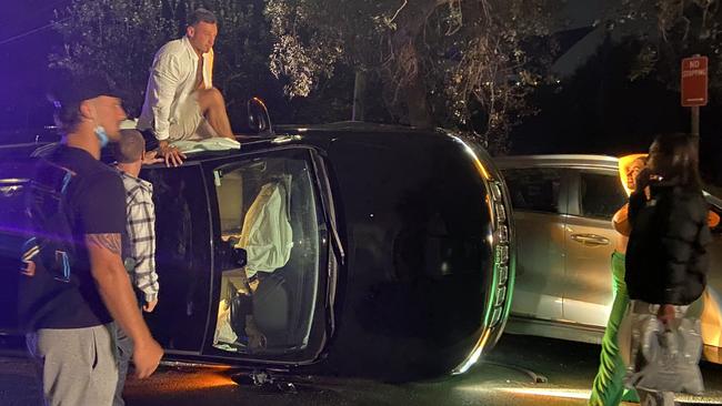 A man with a cigarette in his mouth climbs out of the overturned car, as Ms Hadjia (right) watches on. Picture: Supplied