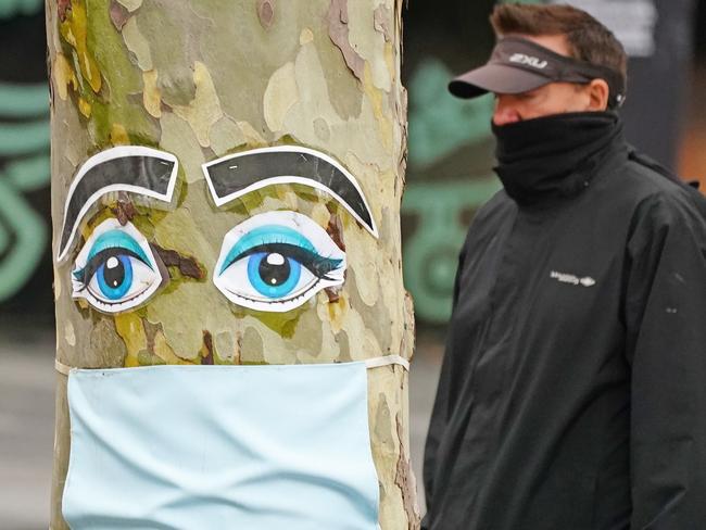 A man walks past a tree with a face mask and eyes stapled to it in Melbourne, Sunday, April 26, 2020. Authorities have said social distancing and isolation measures could still remain in place for the next six months.  A shutdown of non-essential services is in effect Australia wide in a bid to slow the spread of the coronavirus (COVID-19) disease.  (AAP Image/Scott Barbour) NO ARCHIVIN2