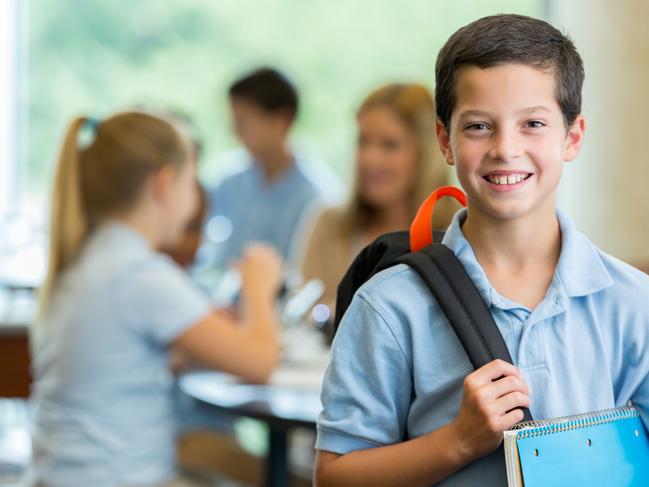 Caucasian male elementary school student smiles at the camera in his classroom. His classmates and teacher are in the background.