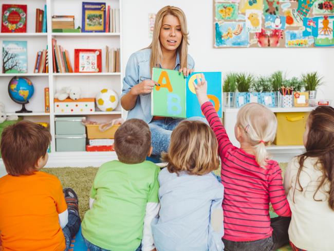 Teacher with a group of preschool children in a nursery. The children are sitting on the floor and listening  teacher. Learning letters. In the background we can see a shelf with some, toys, black board and books.