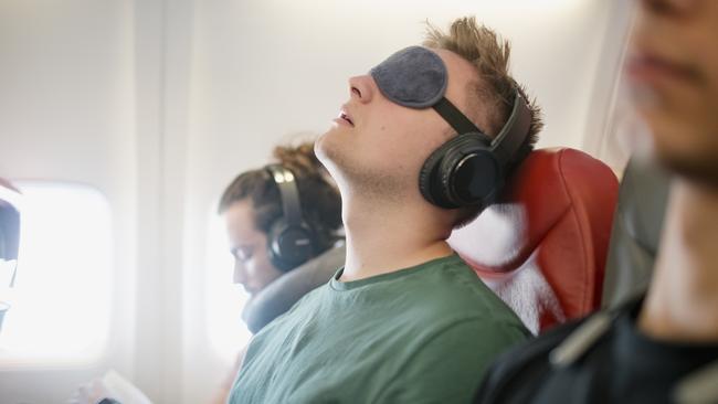 ESCAPE:  Close up shot of a young man asleep with his head back snoring on a flight in economy class. He is wearing wireless headphones and has an eye mask on.  Picture: Istock