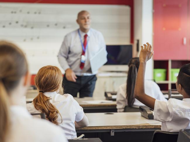 A rear-view shot of teenage music students in a classroom learning how to play the keyboard with their teacher at a school. The students are wearing school uniforms and the teacher is smartly dressed.