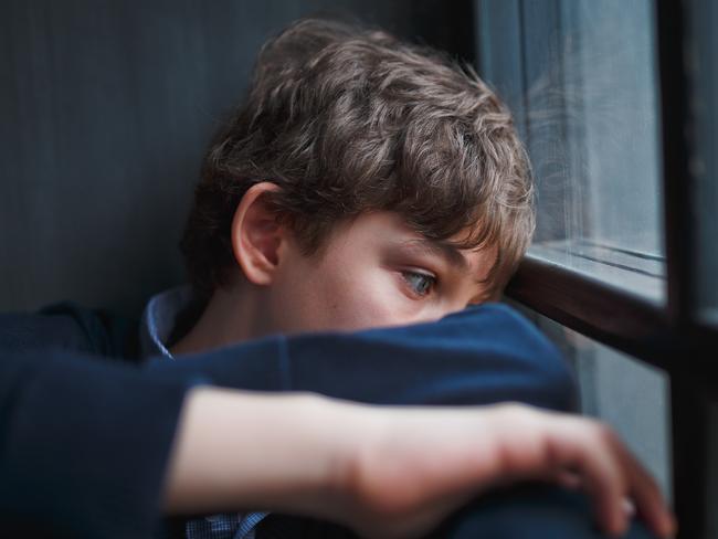 Pensive sad boy teenager with blue eyes in a blue shirt and jeans sitting at the window and closes his face with his hands.