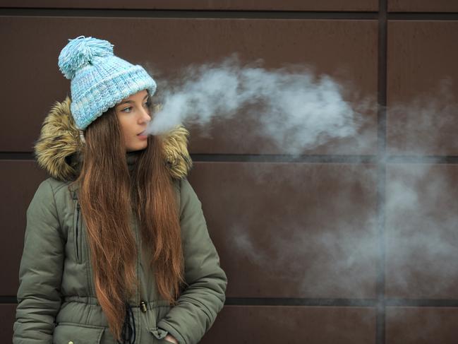 Vape teenager. Young pretty white girl in a cap smoking an electronic cigarette opposite modern brown background on the street in the winter. Bad habit. Close up.