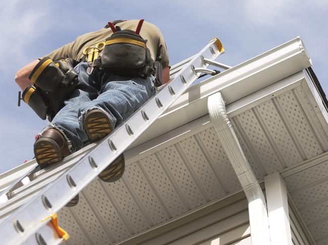 A handyman repairs his rain gutters.  He is up a ladder, photo taken from ground looking up, low angle view.  He wears a tool belt, sky and clouds, good copy space. tradesman generic